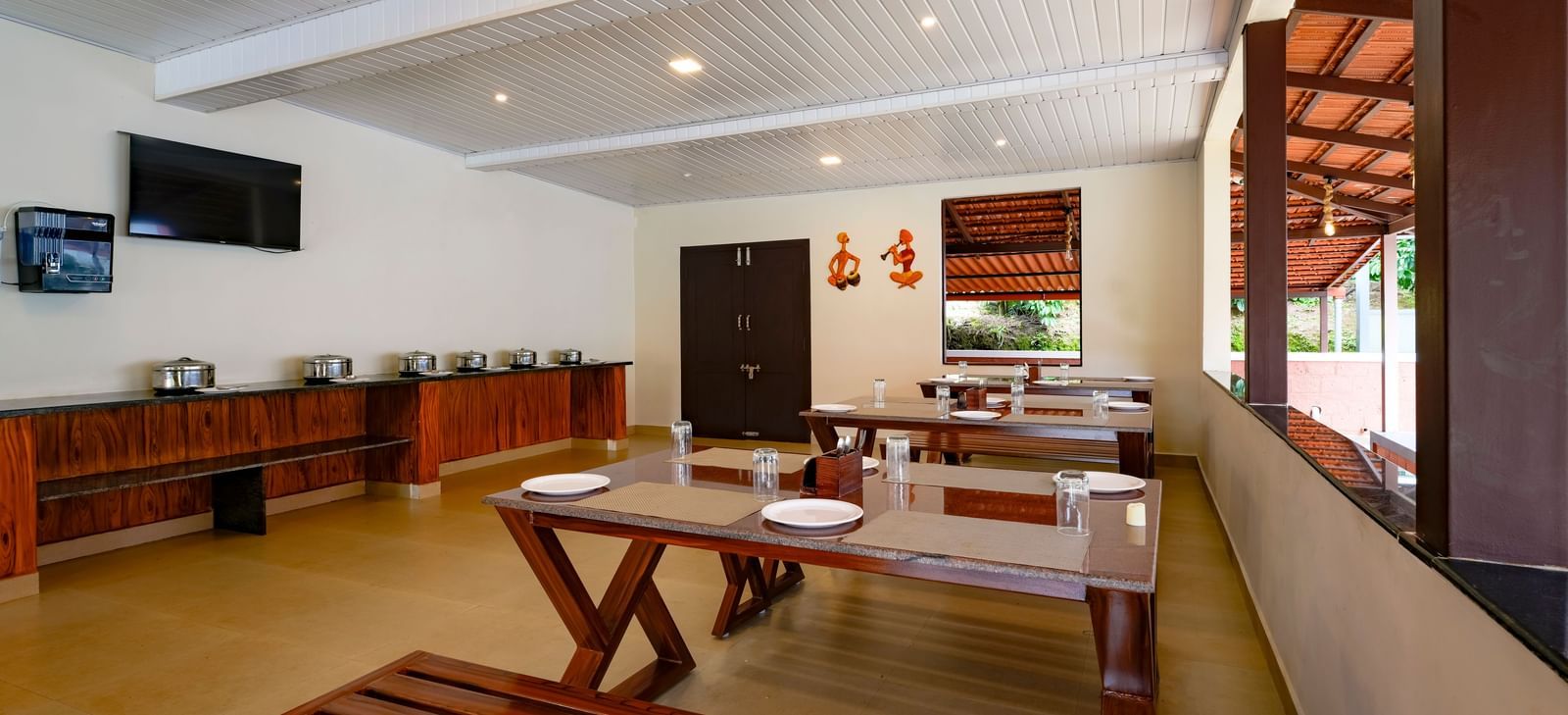 A daytime interior view of the Mane Cafe at Bhuthanakadu Retreat, highlighting a buffet area with covered dishes, a TV on the wall, and long wooden dining tables with benches.