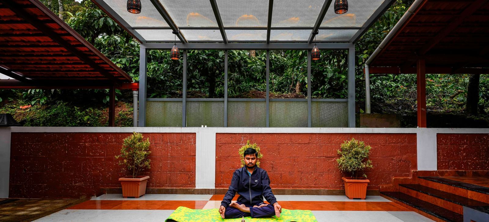 An individual is meditating in a serene, partially covered outdoor yoga space at Bhuthanakadu Retreat, surrounded by lush greenery and red-tiled walls.