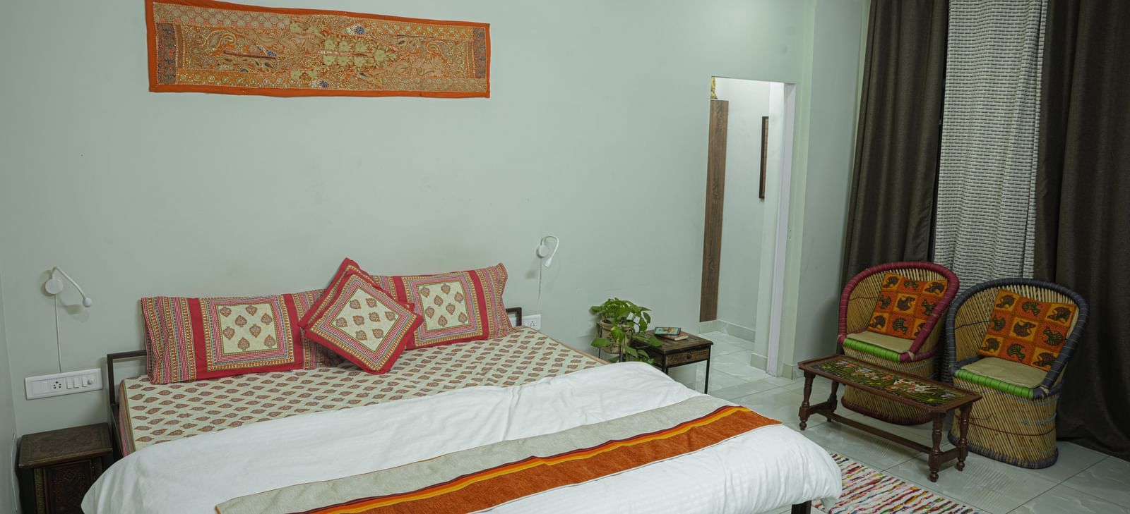 A well-kept bedroom with a double bed, white bedding, orange throw, chairs and wall decor at Jaypore360 Homestay - Best Rooms in Jaipur.
