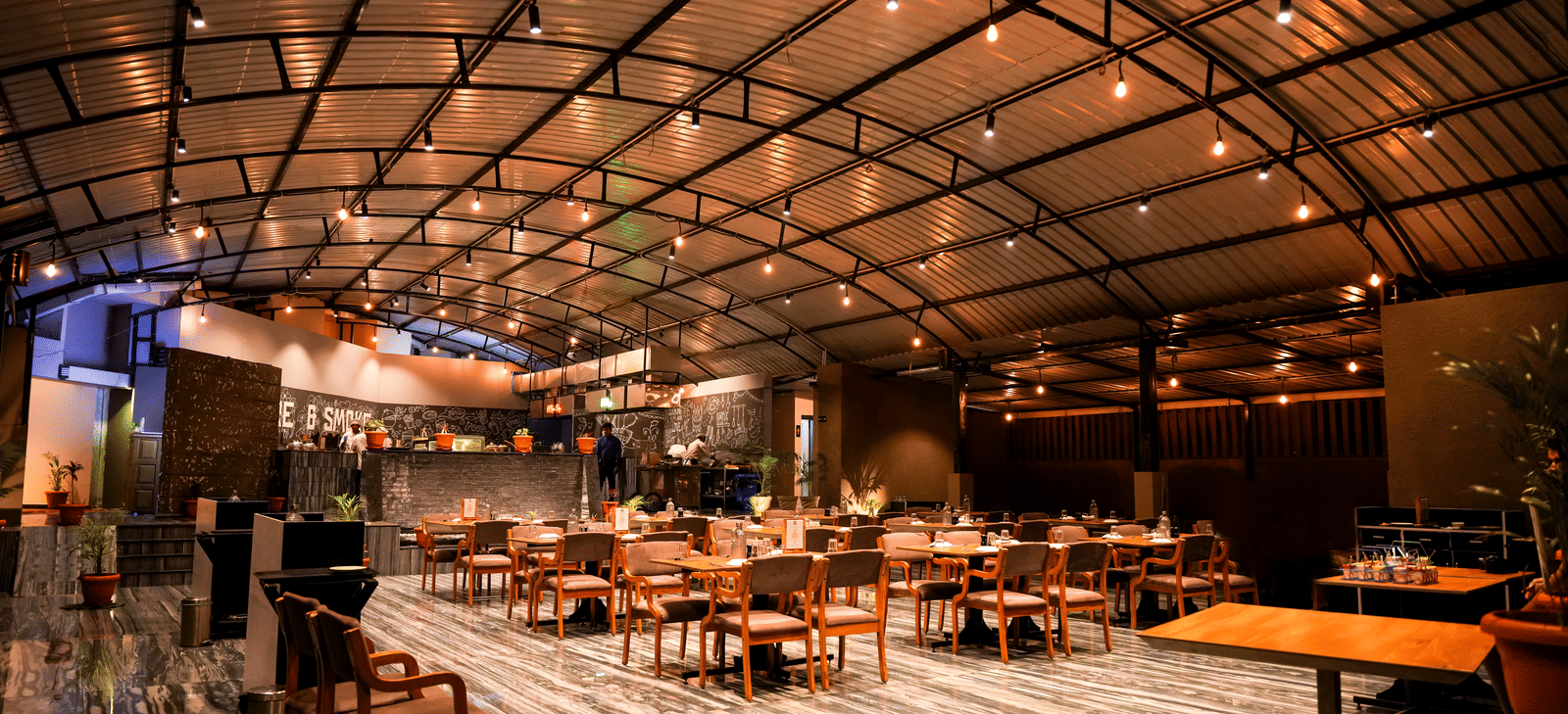  A wide view of the spacious dining hall with wooden tables and chairs under a curved, industrial-style roof at Pravasa Hotel, Kolhapur.