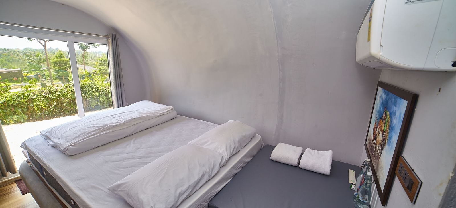 The interior of a modern, capsule-style accommodation unit featuring two single beds with white mattresses and a small bedside table, with a window offering a view of greenery. - Vintage Trails by Ilara