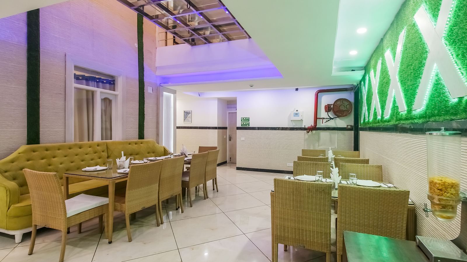 A restaurant interior with a long tufted sofa, wooden tables and chairs, a decorative wall design, and a tiled floor, lit by soft lighting. - Hotel Diamond Plaza, Chandigarh.