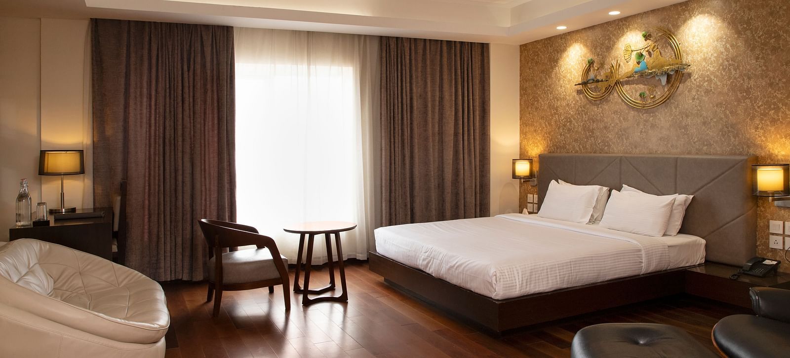 A spacious room featuring a large bed, a cosy seating area, elegant curtains, soft ambient lighting, and expansive windows with curtains drawn to the side at The Hamuse Hotel, Kodaikanal.