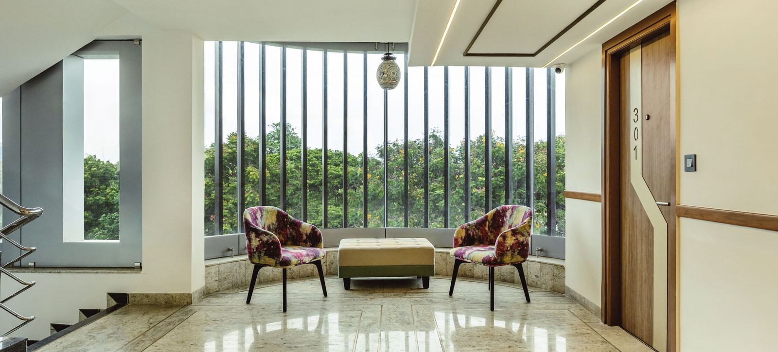 A seating area with a sofa and two chairs at Mango Hill Central, Chennai. Large windows with a view of greenery are visible.