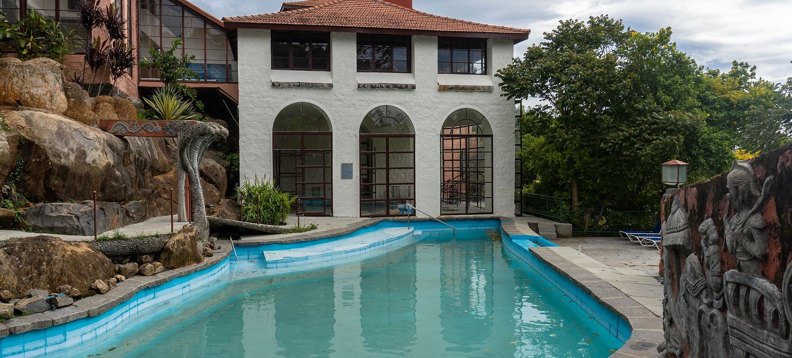 A white building with arched windows and a red roof next to a blue swimming pool at Mango Hill, Yercaud.