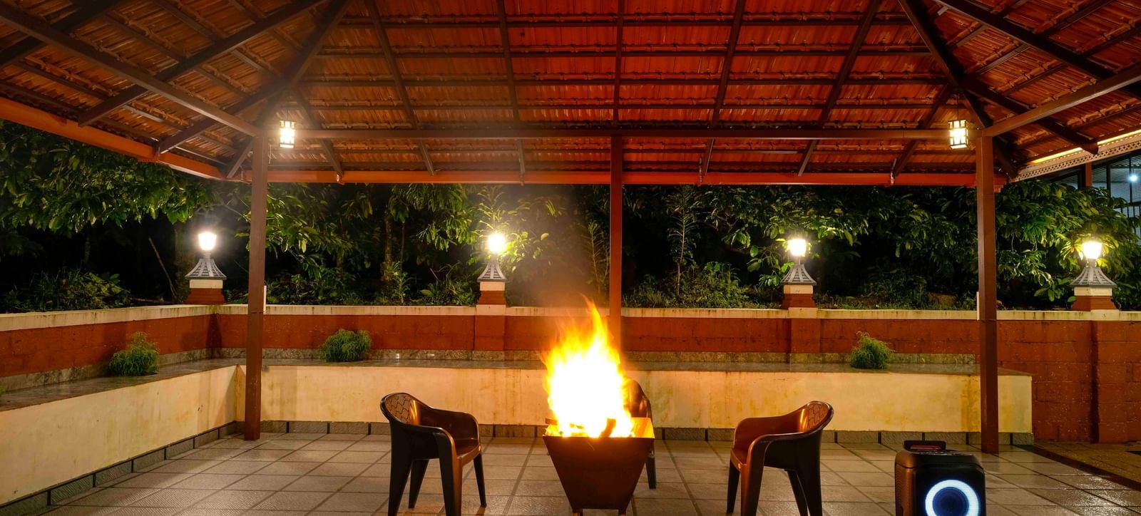 A large bonfire crackles at the center of a covered, open-air pavilion at Bhuthanakadu Retreat, surrounded by chairs under the warm glow of the lights at night.
