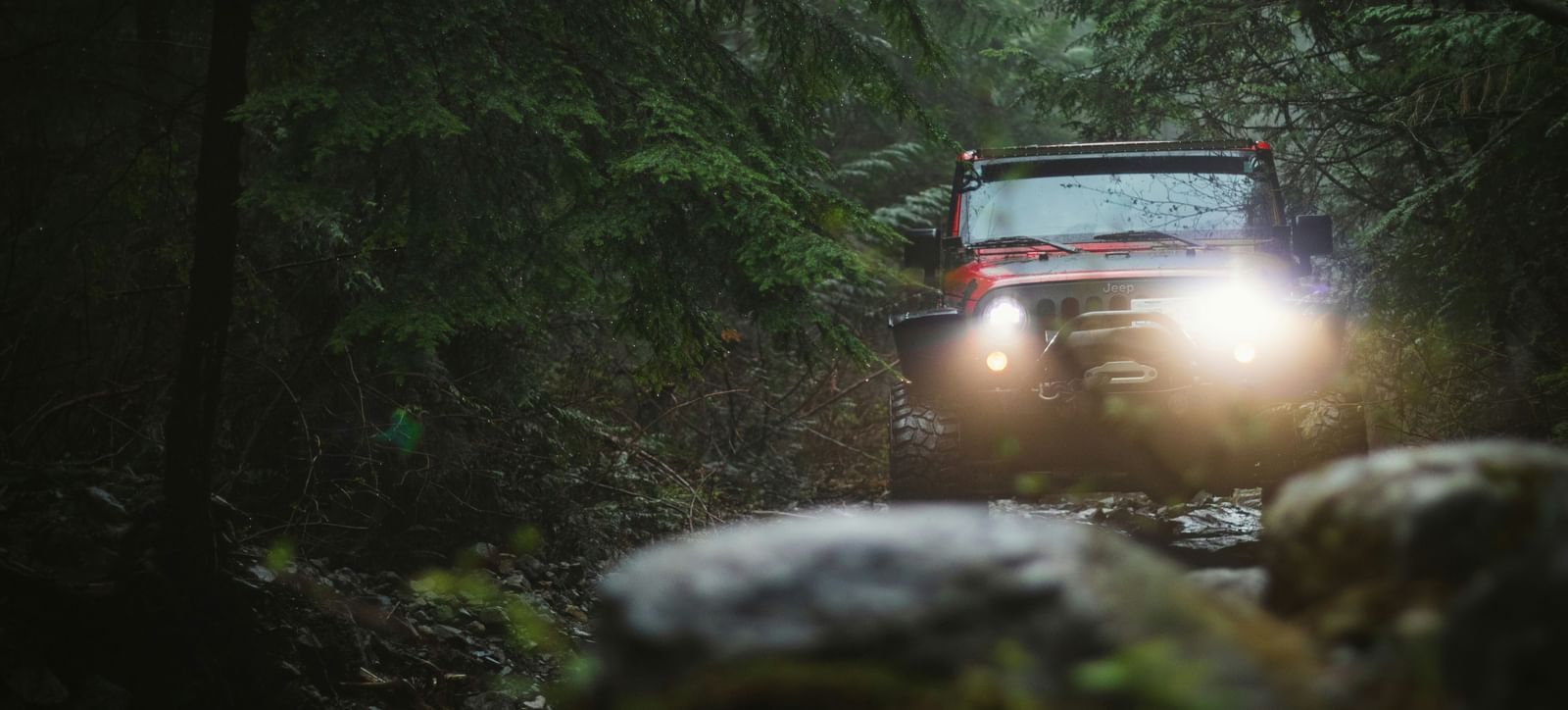 A red jeep with headlights on drives down a rocky, wooded path surrounded by dense green trees.