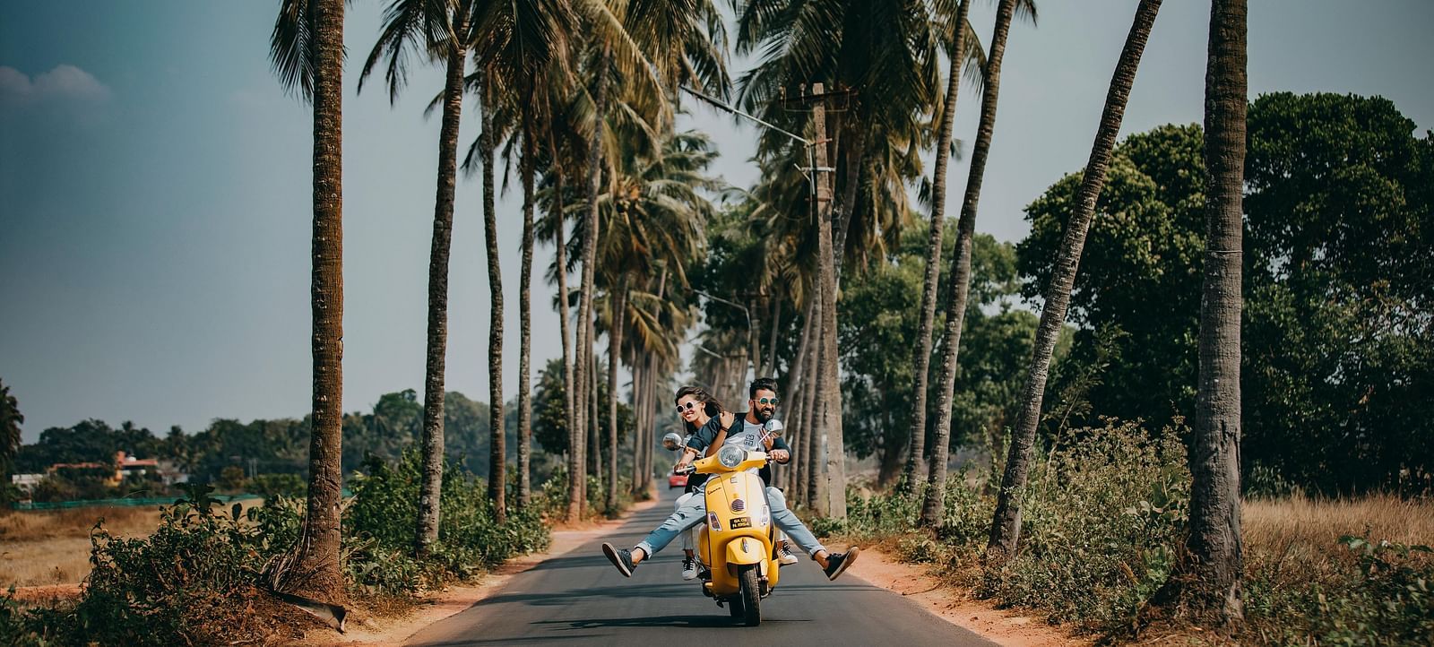 A person rides a yellow scooter down a paved road lined with tall palm trees under a bright sky.