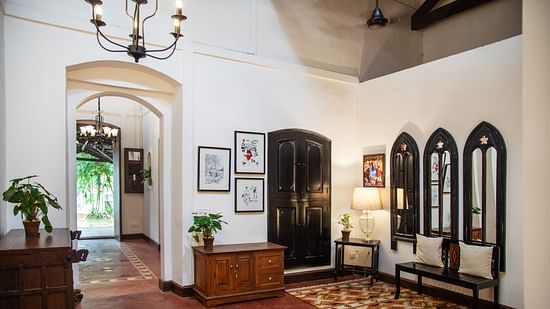 An image of a huge lobby where various decorative items are placed on top of the cabinets and shelves, there is another table with lamp and three vintage style window near it - Lamrin Ucassaim Hotel, Goa