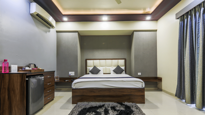 A modern hotel room with a double bed, padded headboard, side tables, mini-fridge unit with kettle, wall AC, ceiling lights, patterned rug, and floor-length curtains along the right side - Magnus Vosiv Suites, Kalyani Nagar