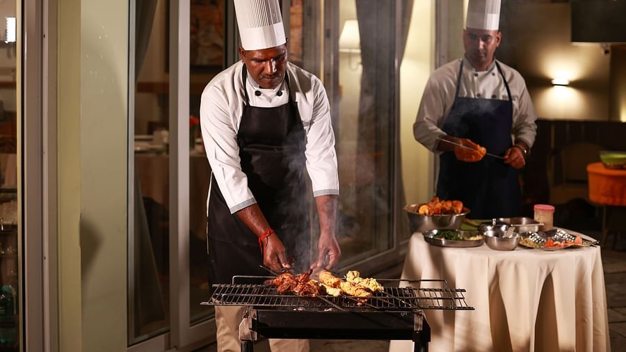 a chef grilling food on a barbecue while another chef can be seen prepping the food in the background - Lamrin Norwood Green, Palampur