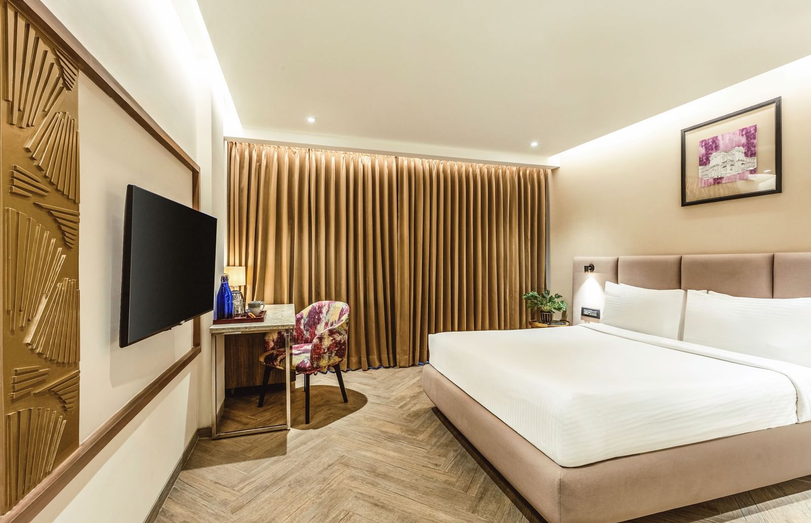 A wide-angle view of a room with a large bed and a television at Mango Hill Central, Chennai. A desk and chair are visible on the right.