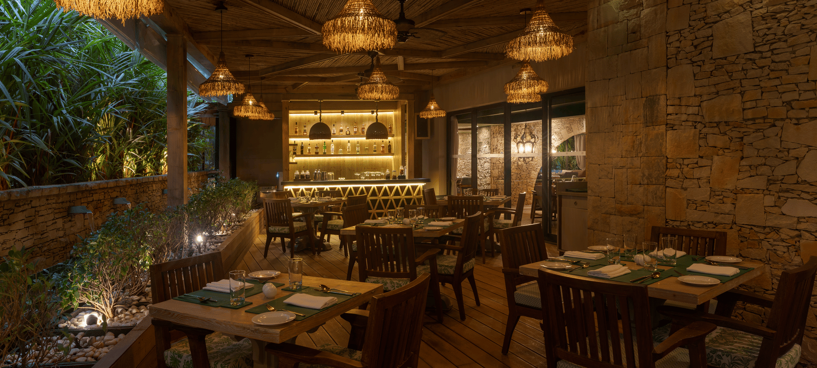 A dimly-lit dining area at the Mila Restaurant with lush green trees on one side at The Evren, Vagator.