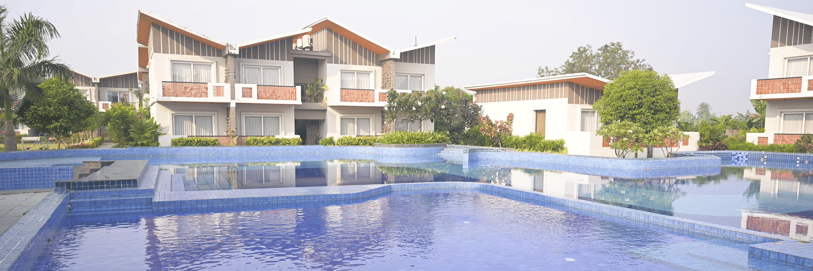 A calm swimming pool reflecting nearby villas and greenery on a sunny day at Hotel Sonar Bangla Mayapur.