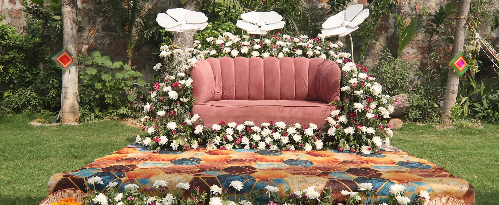 Flower-decorated wedding stage at Tisya Farms, Gurgaon with cushioned seating and white, red, and orange floral arrangements.