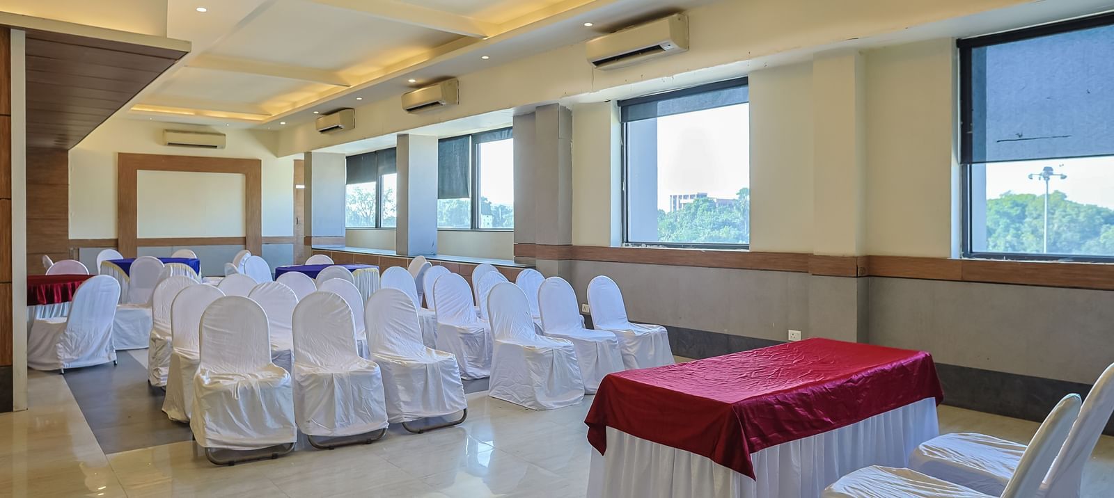 Banquet hall with rectangular tables, white chairs and large windows at Max Hotel Prayagraj.