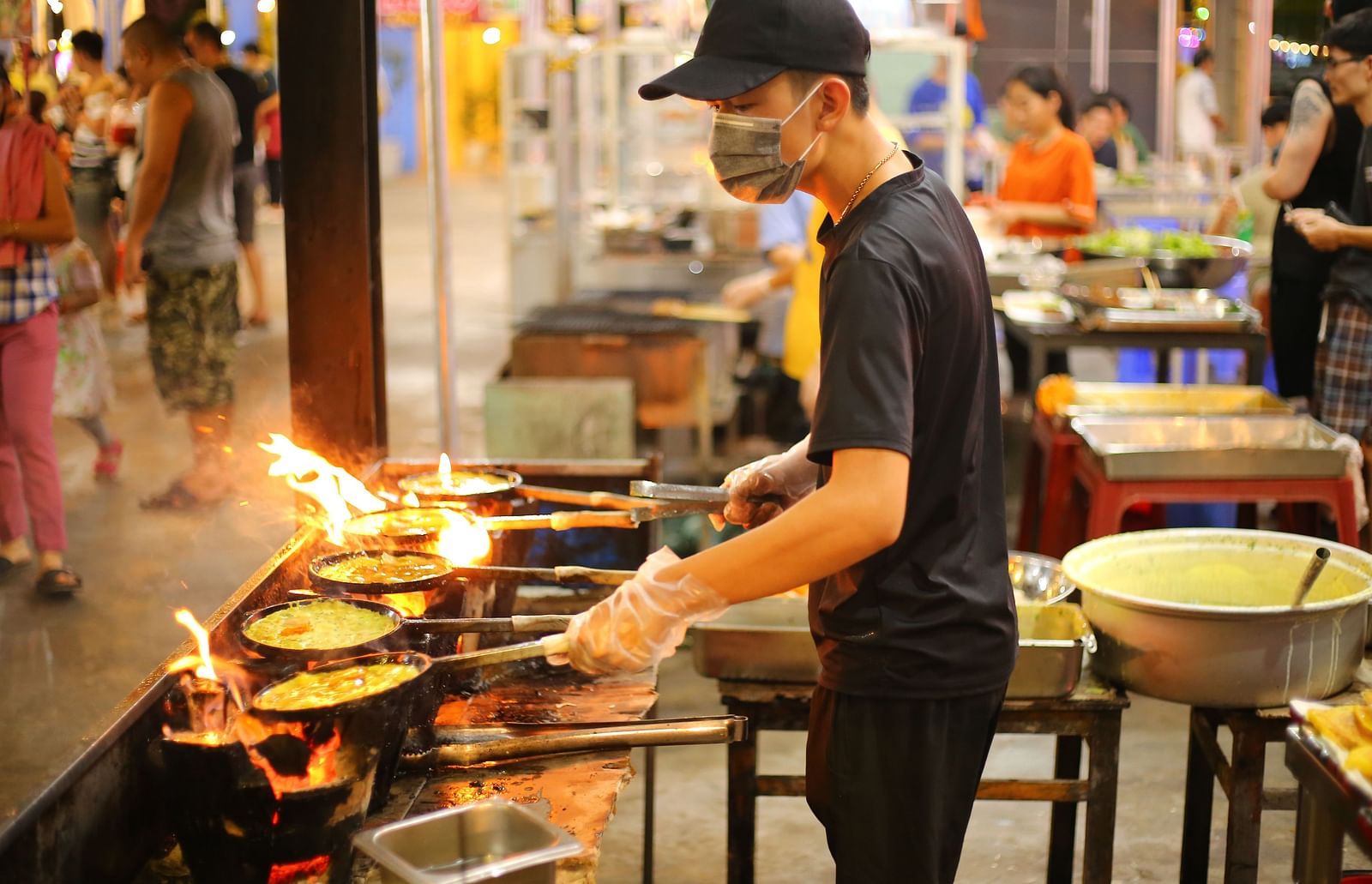 A person cooking on many dishes on the street with other people in the background.