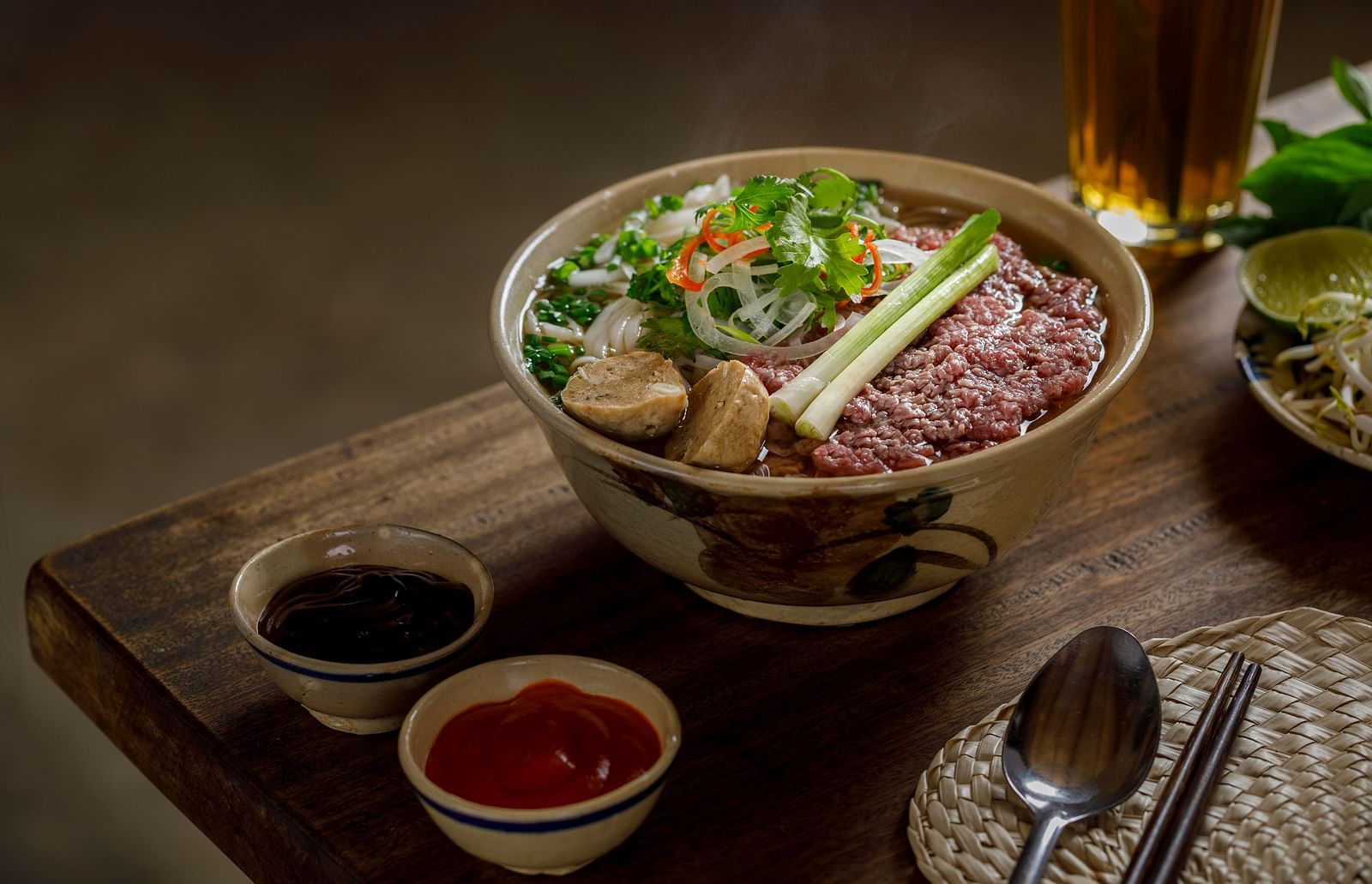 A close up of Pho in a bowl with garnishing on top of it with two sauces next to it.