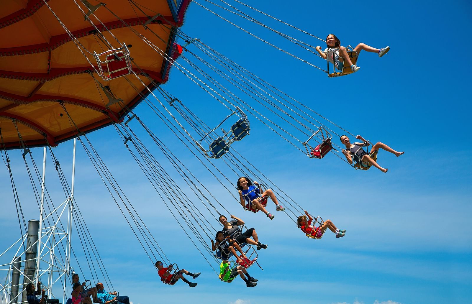 A swing carousel spins high under a bright blue sky, carrying riders in suspended seats. 