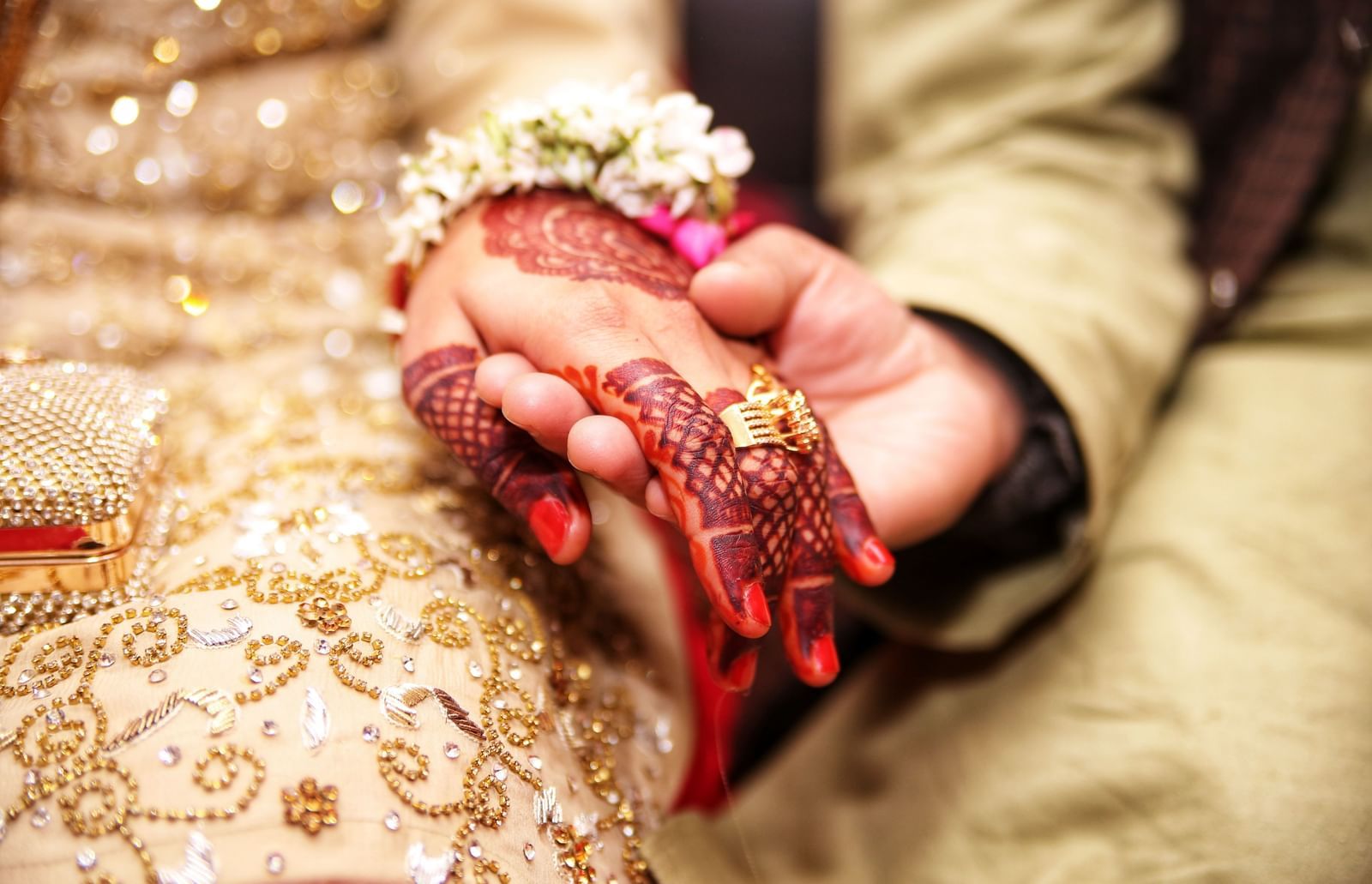 Close-up of a bride and groom's hands clasped together. The bride has elaborate henna on her hand, a gold ring, and a floral wrist ornament. Her gold dress is visible.