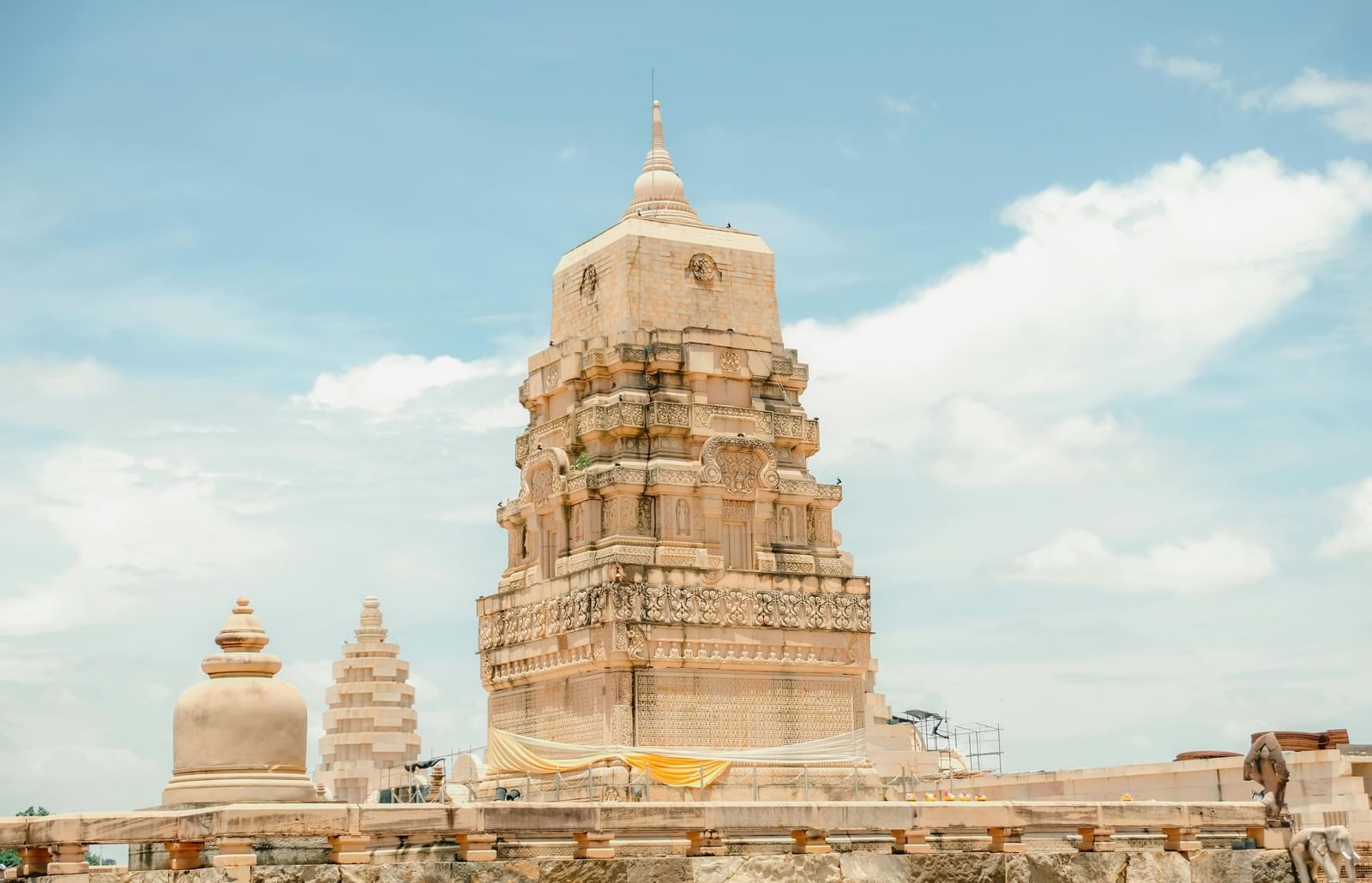 A tall, white temple with a conical tower (shikhara) and several smaller domes, captured from a low angle against a cloudy blue sky.