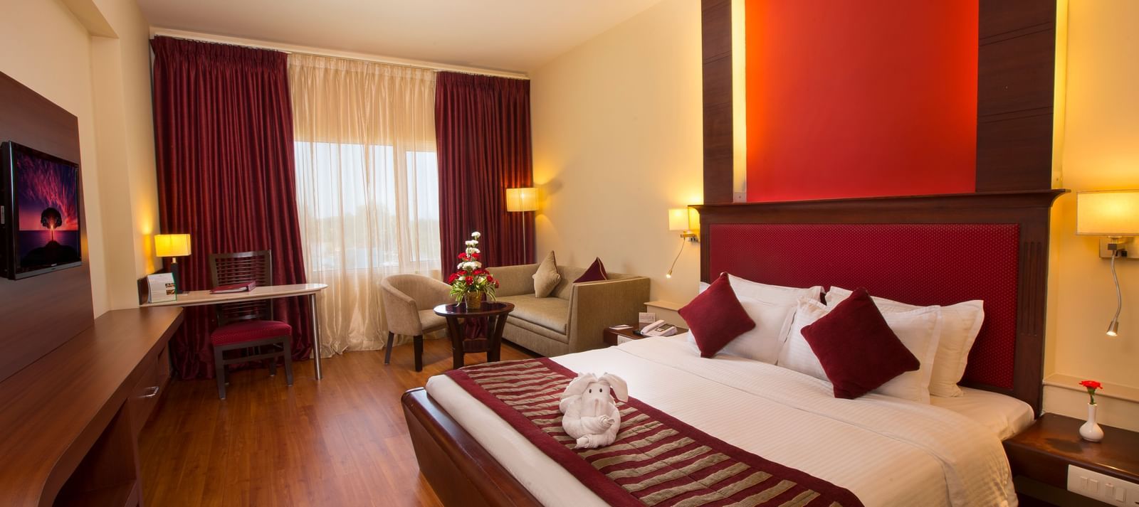 Bedroom with a rabbit made with towel on a large bed, table, chair and curtained window at  Hotel Atithi TGI Grand.