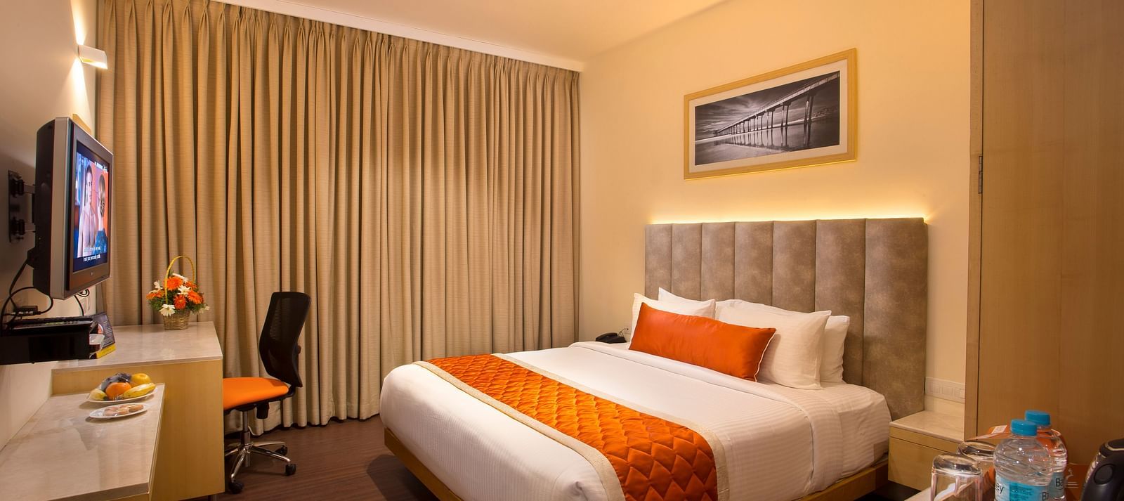 A cosy hotel room with a plush bed adorned with an orange bed runner, a work desk and a wall-mounted TV - Hotel Langford Keys by TGI, Shanti Nagar