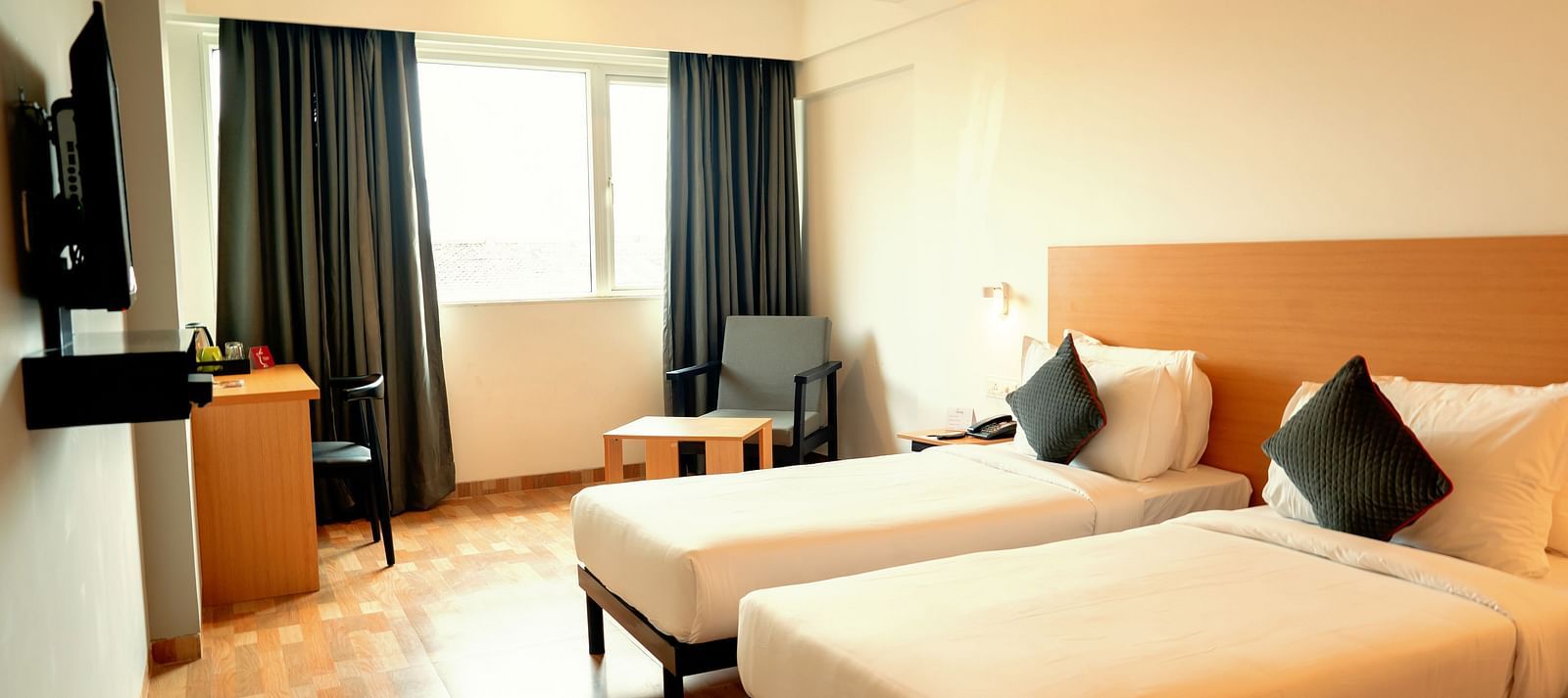A bright hotel room with two single beds, a desk and a flat-screen TV, with a large window draped in gray curtains - TGI Ameya Grand, Goa