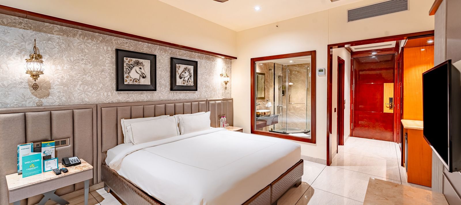 A spacious bed with two photo frames above, a bedside table with essentials, and a wall mounted TV opposite at  Crescent Spa & Resort, Indore