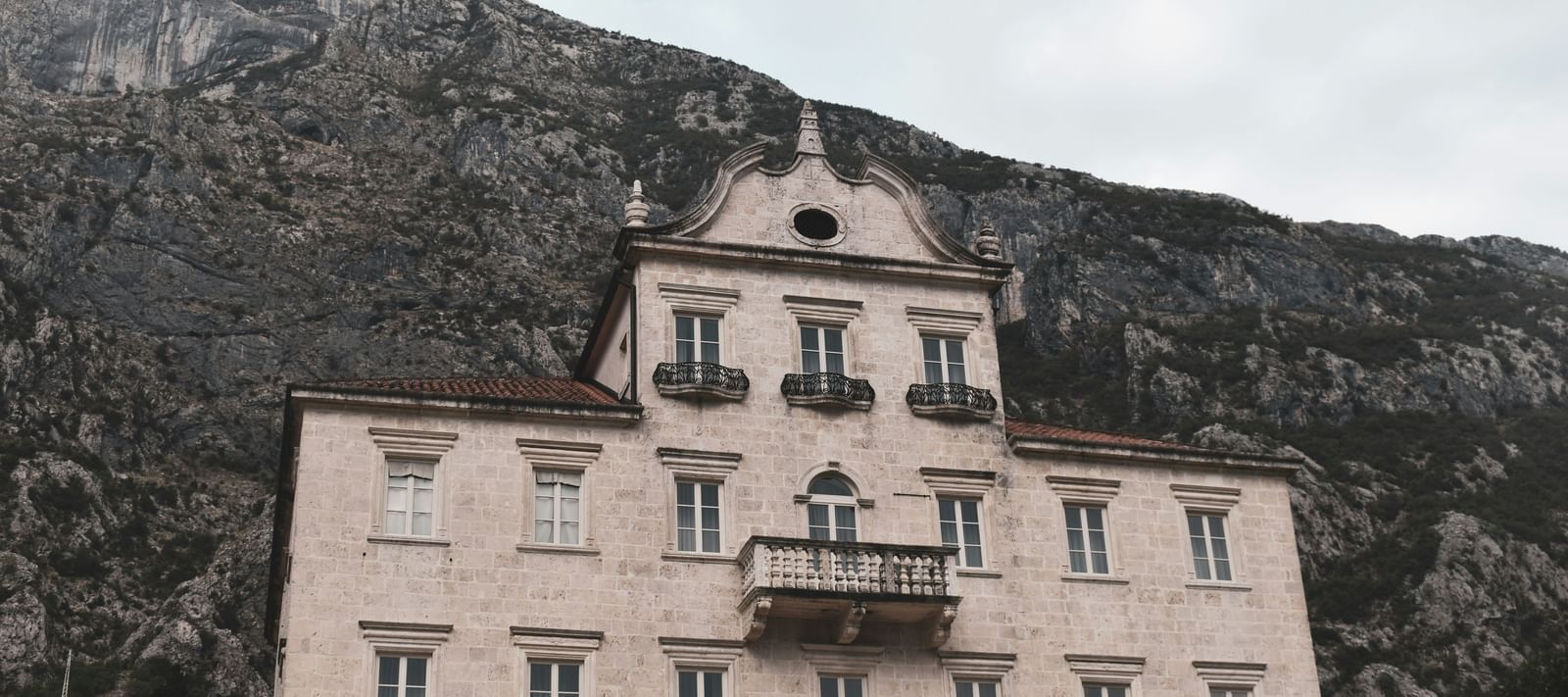 An old, majestic, multi-story building with a balcony and an arched entryway, set against a backdrop of a large mountain.