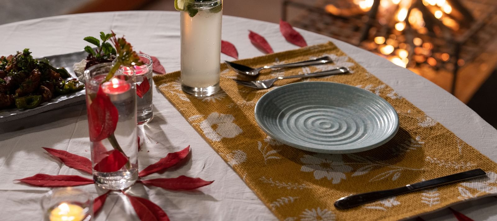 A close-up view of a table neatly set with food and cutlery, with a campfire in the background at BluSalzz Villas - The Green Roof, Bhimtal.