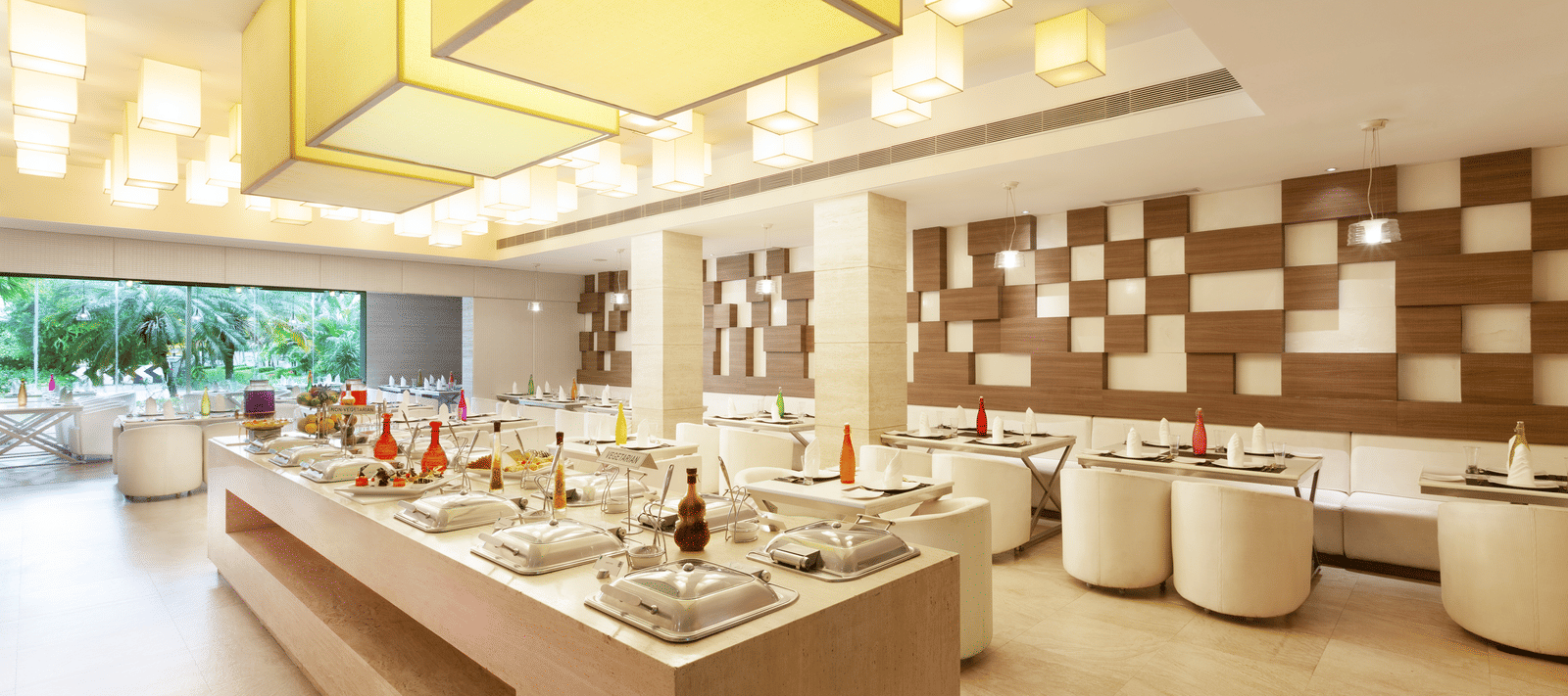 A diagonal view of a restaurant with an island in the middle featuring buffet items, surrounded by tables and chairs on the sides | Cenneys Gateway