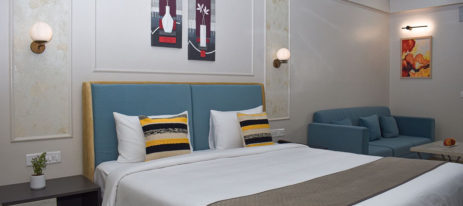 A hotel room with a bed, a blue sofa, a small table, wall-mounted light fixtures, and framed artworks - Urban Suites by BluSalzz, Jaipur