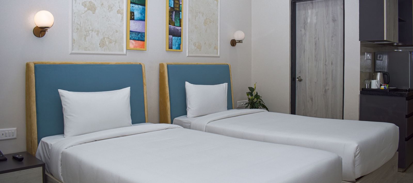 A hotel room with two twin beds, wall-mounted light fixtures, framed artwork, and a partial view of a kitchenette - Urban Suites by BluSalzz, Jaipur