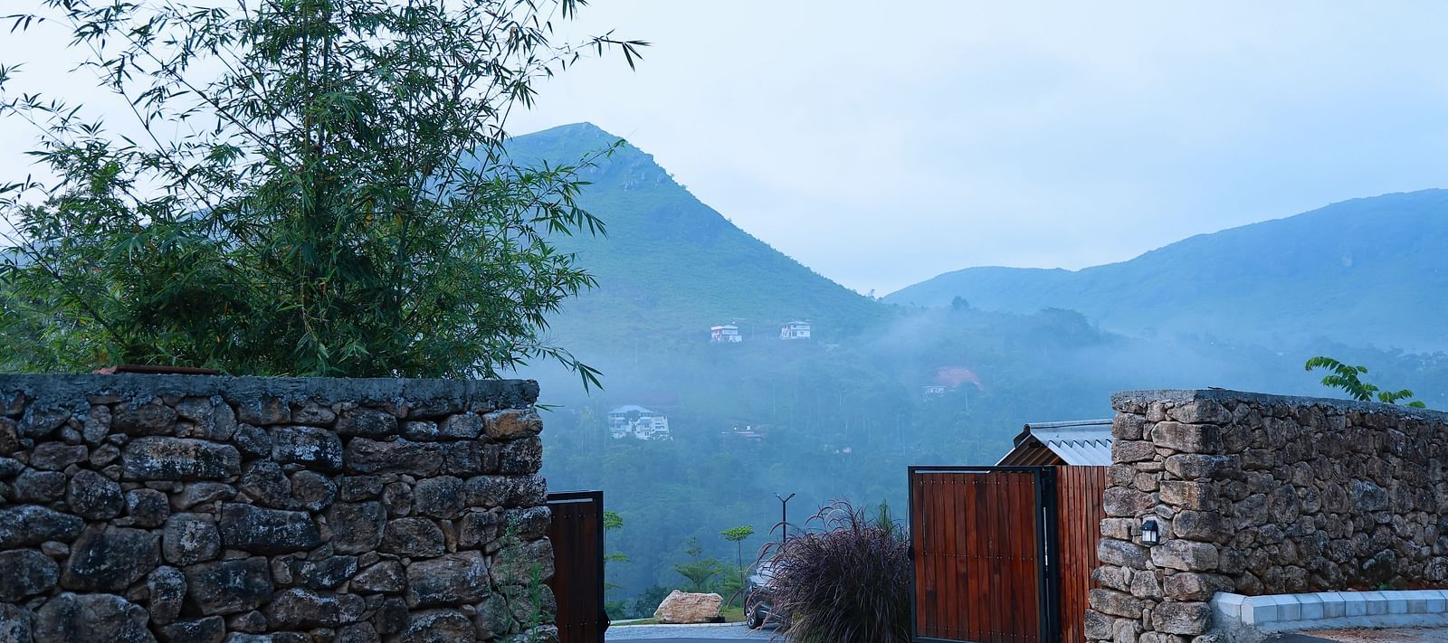 A stone wall entrance with an open wooden gate at Vaasta Vagamon, revealing a winding road and distant mist-shrouded hills.