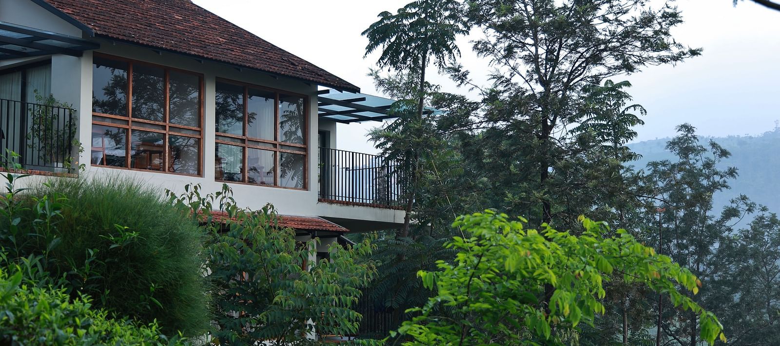  A building at Vaastha Vagamon, nestled amidst abundant foliage, with large windows overlooking a verdant landscape.