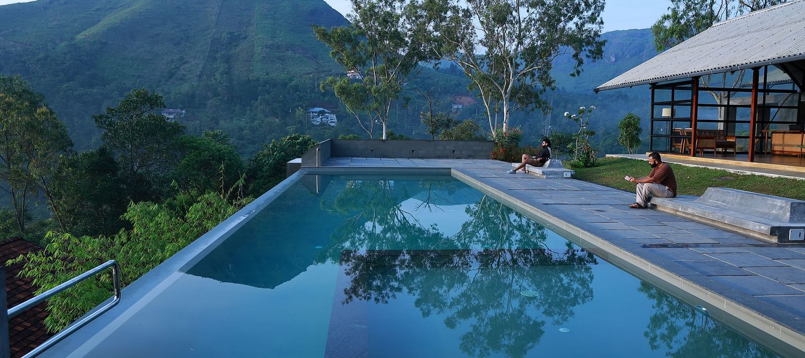 An infinity pool at Vaastha Vagamon overlooking a serene mountainous landscape, with guests relaxing on the poolside deck.