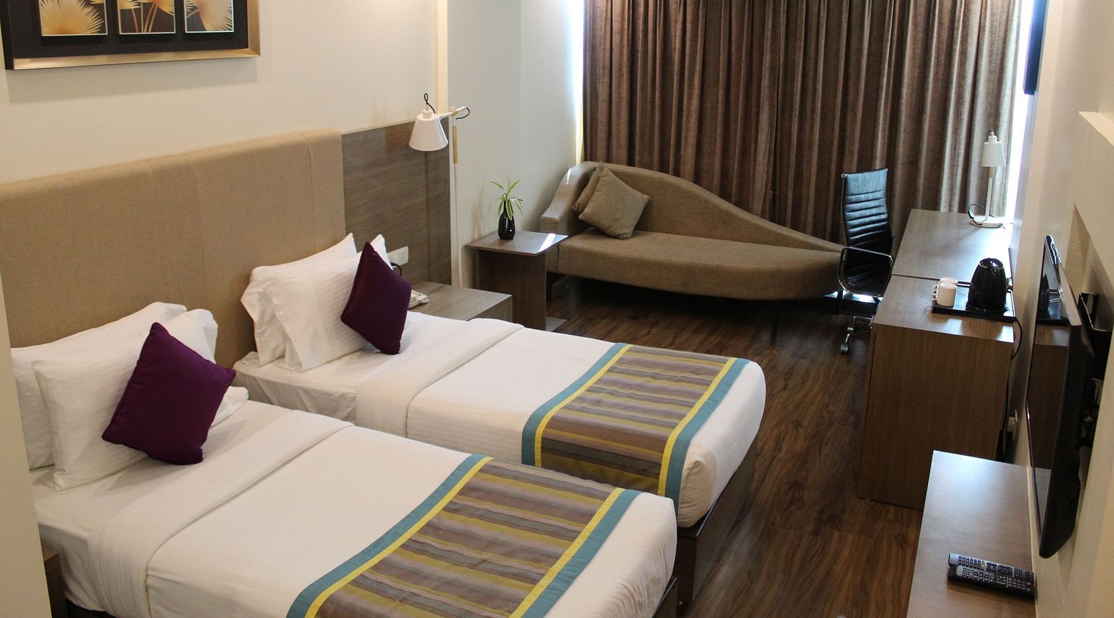 An image showing a pair of twin beds with lush decors, a long sofa with cushion, and a flat screen TV, inside a room with closed curtains.- Golden Suites, Gurgaon