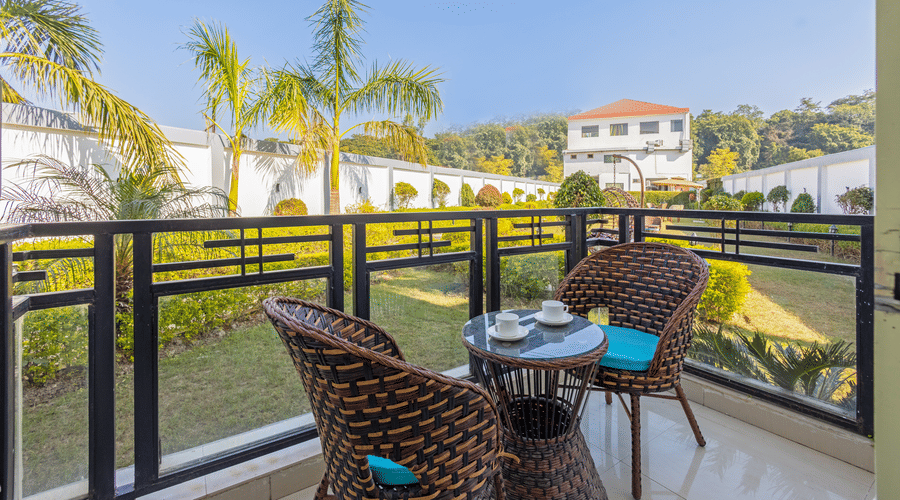 A sunny balcony with two wicker chairs and a small table overlooking a lush green garden - Nature Trails Rishikesh