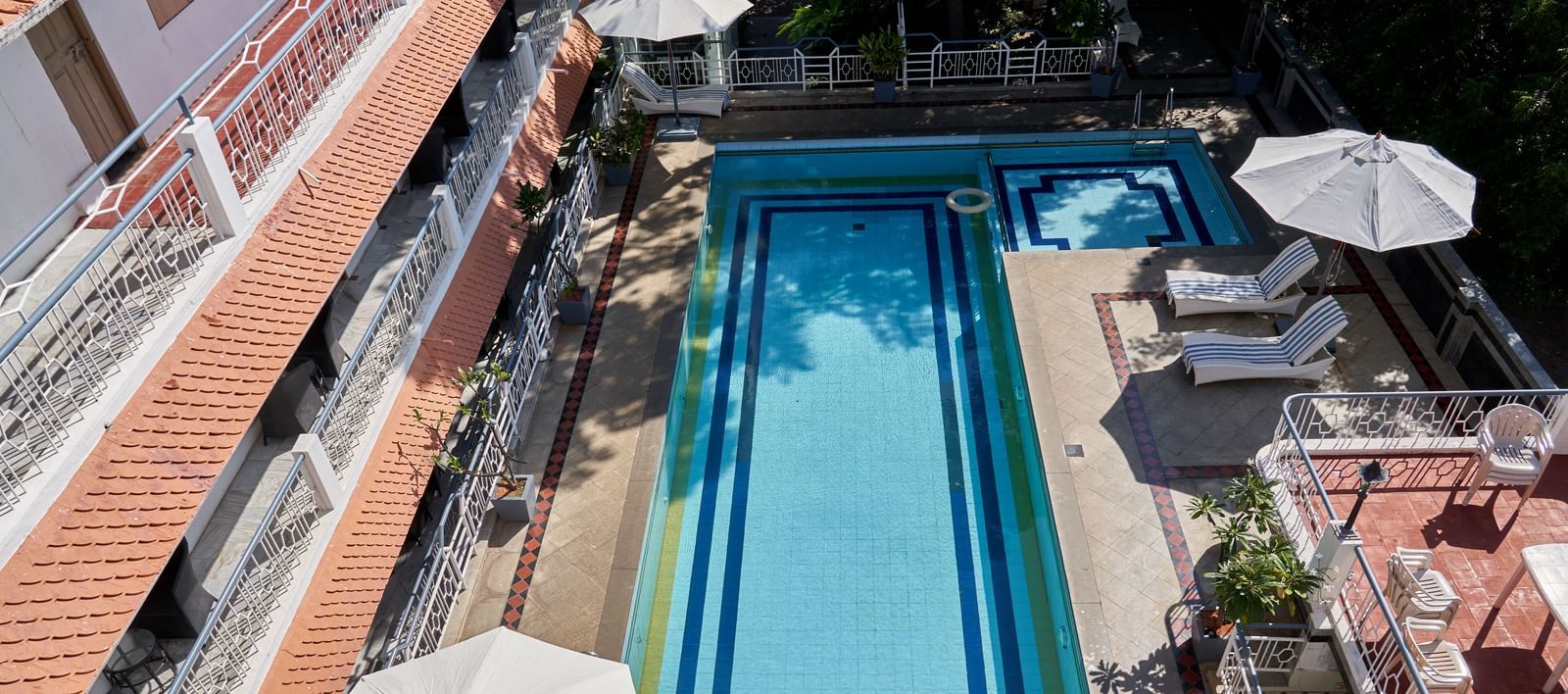 A slightly elevated view of a long, rectangular swimming pool at Hotel Mamallaa Heritage, with white umbrellas and the hotel building visible beside it.