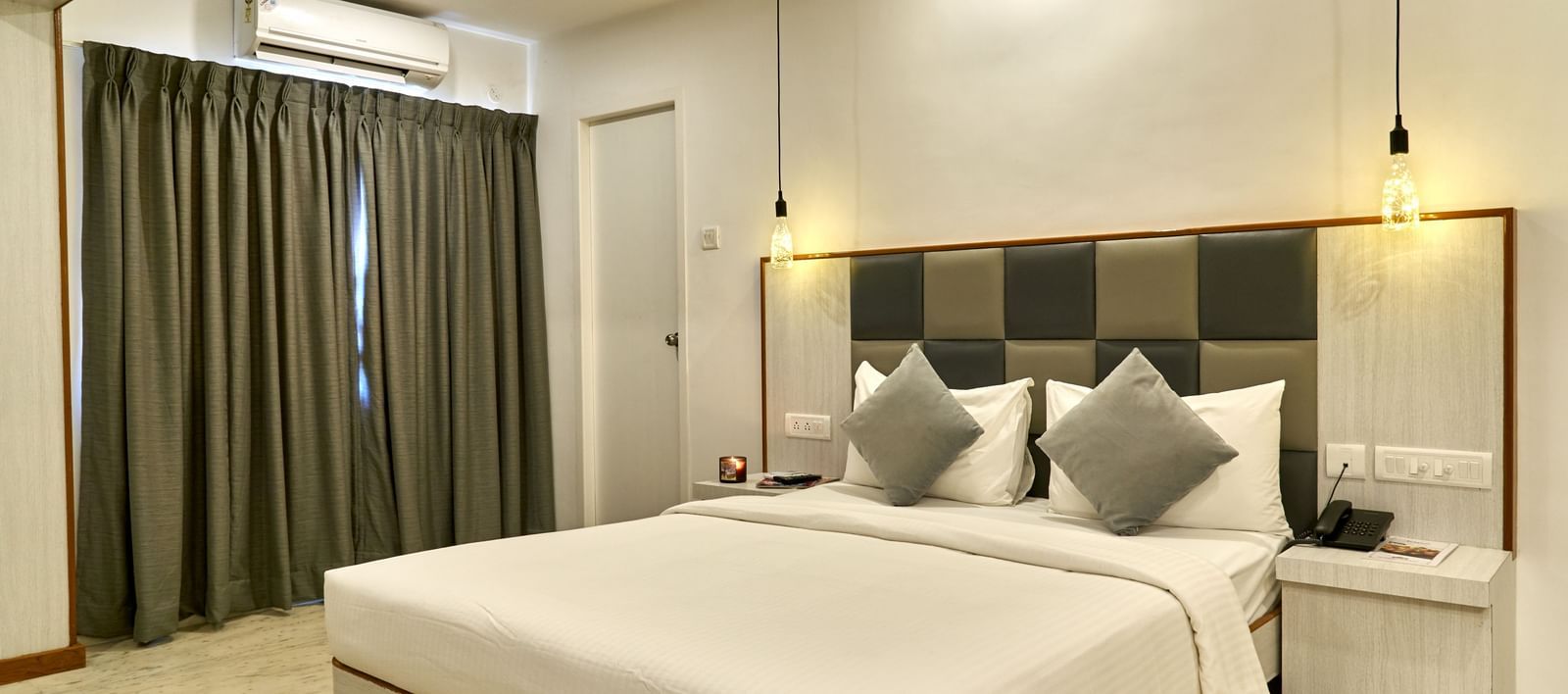 A clean and contemporary hotel room at Hotel Mamallaa Heritage, showcasing a bed with a light-coloured headboard, crisp white bedding, and modern lighting fixtures.
