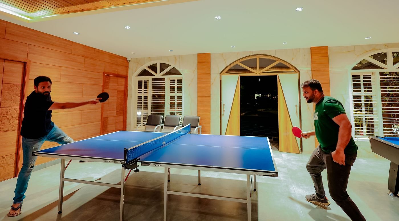 Two people play ping pong in a room with brick walls, large windows, and a high ceiling with a decorative light fixture - Nirantara Resort & Spa.