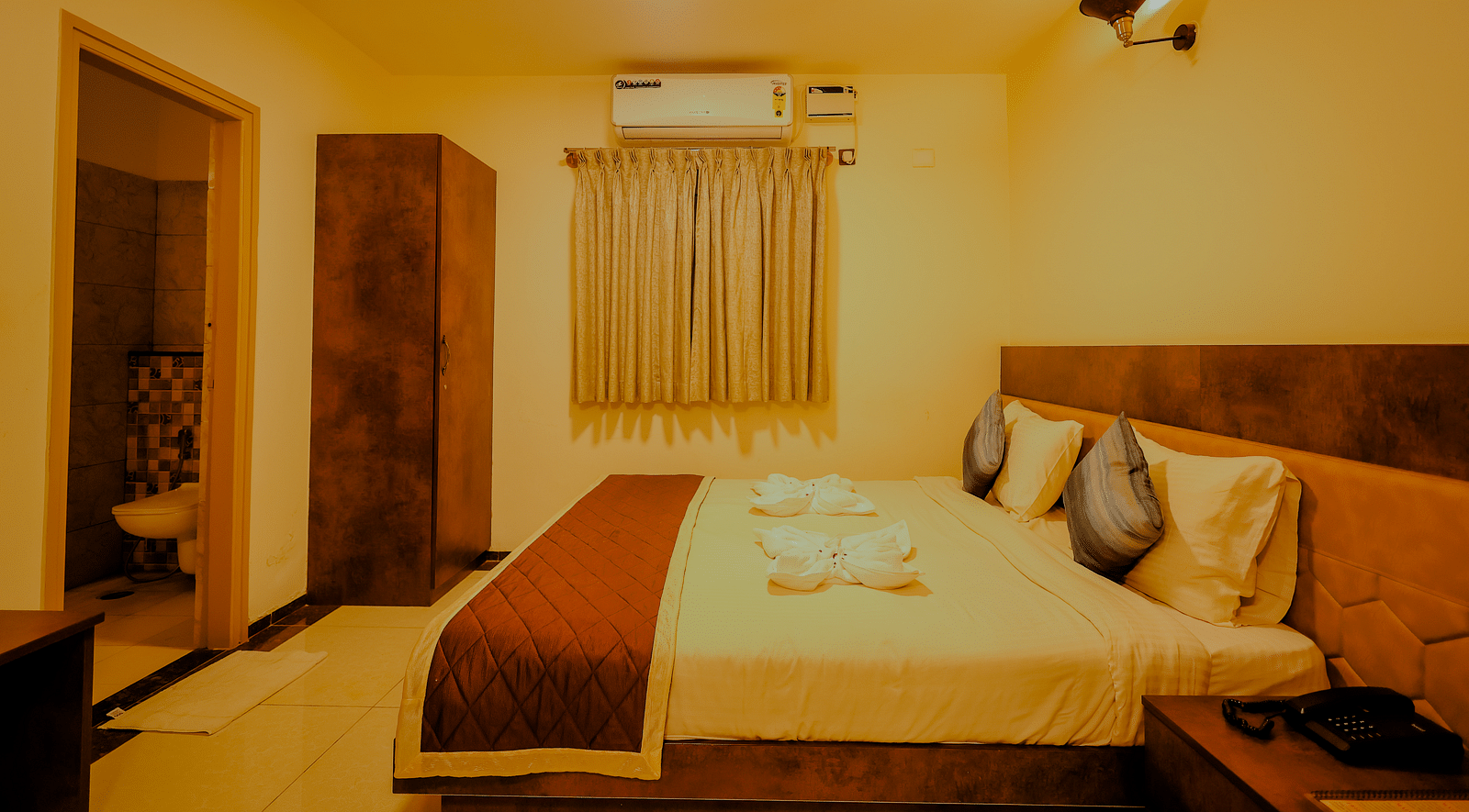Side angle view of a room at Niratara Resort and Spa featuring a bed, window with short curtains, an almirah, and the bathroom door.
