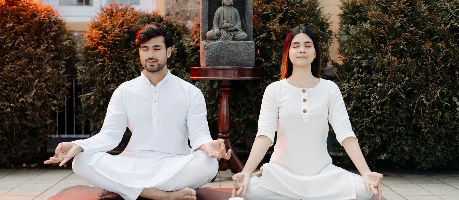 Man and woman meditating together in a serene outdoor space, dressed in white attire symbolizing tranquility.