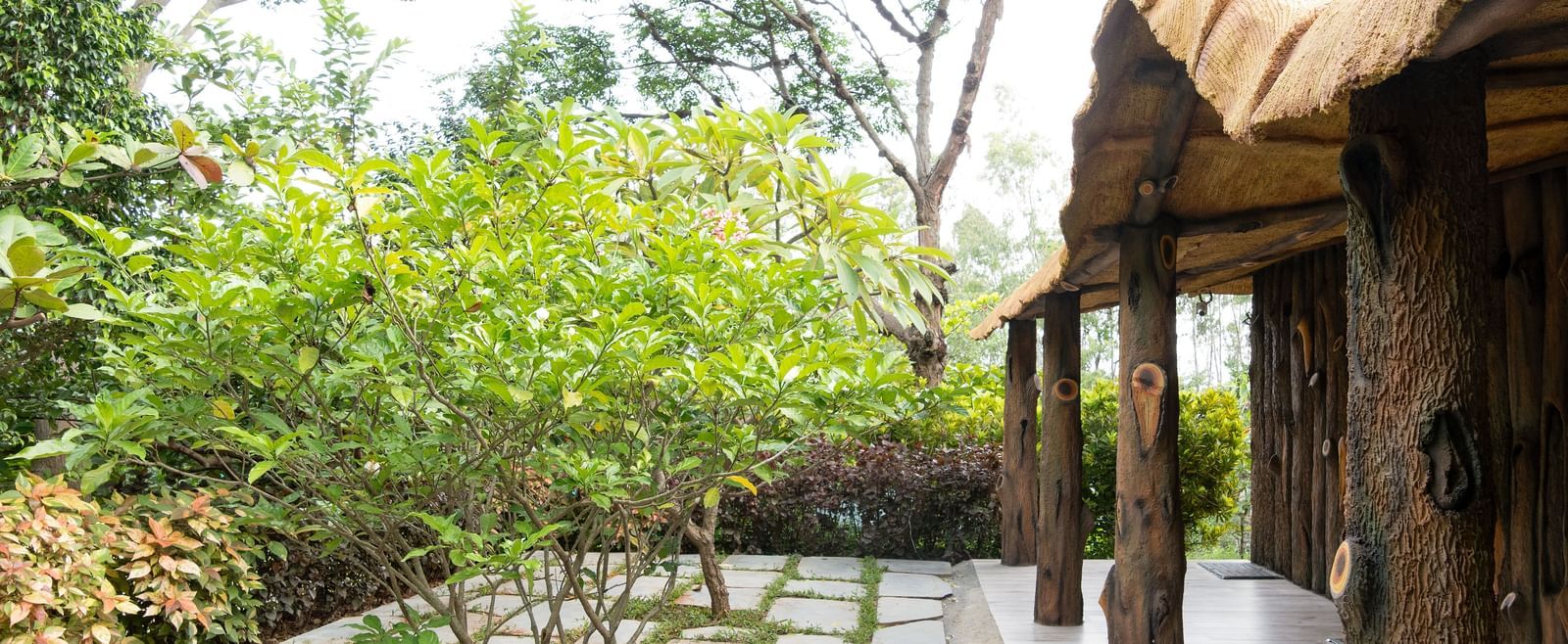 An outdoor pathway with stone pavers is bordered by manicured green bushes and leads towards a building with a thatched roof | Standard Room | RD's Nature Retreat, Bangalore
