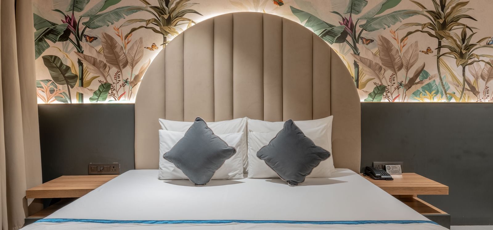 A front-facing view of bed with arched padded headboard and tropical-themed wall mural at Hotel Highway King, Jaipur.
