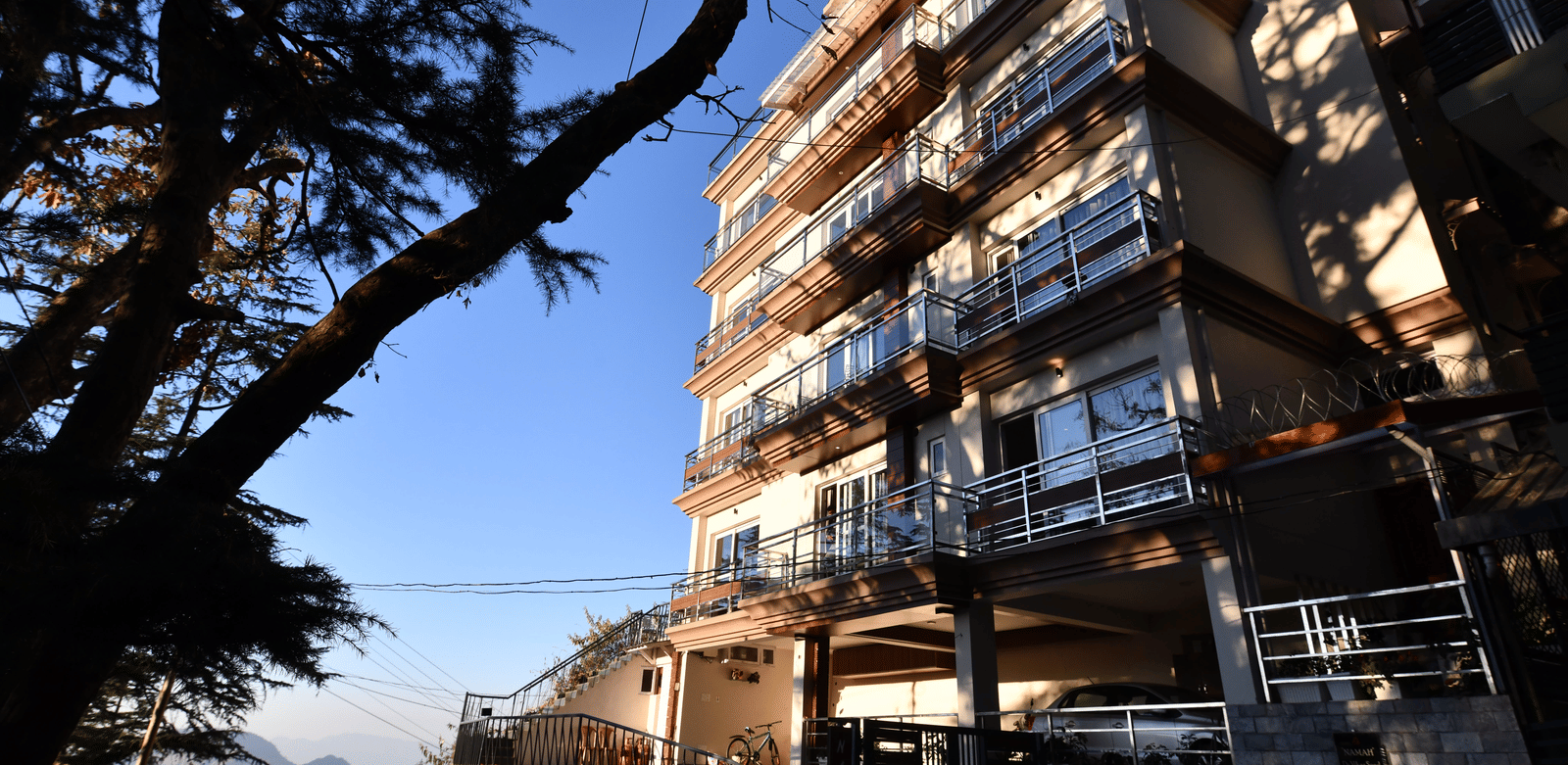 A facade of Perfectstayz Value Shimla (Namah Retreat) with balconies viewed from below against a clear sky and a tree in foreground.