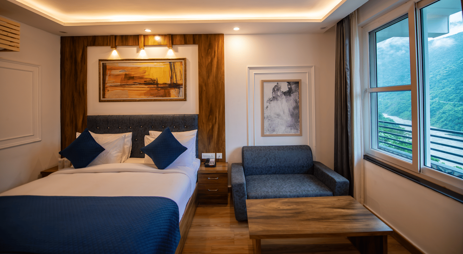 A Deluxe Room at The Tattva Devaprayag with a bed featuring blue blankets and two pillows, two framed paintings on the wall and a sofa.