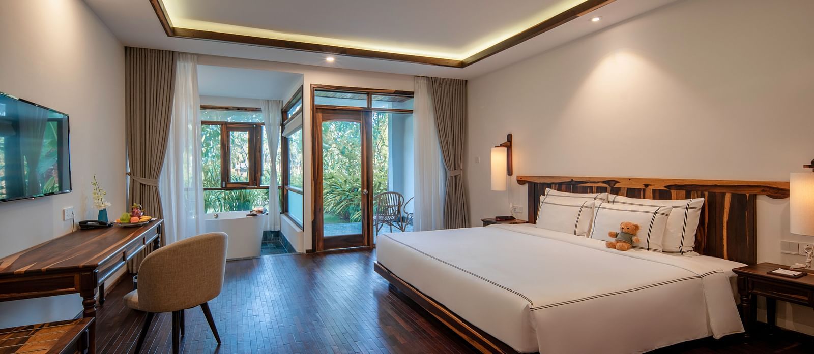 A view of a room with a wall mounted Tv in front of a large bed and a glass door opening to a garden - Albu Resort Nha Trang
