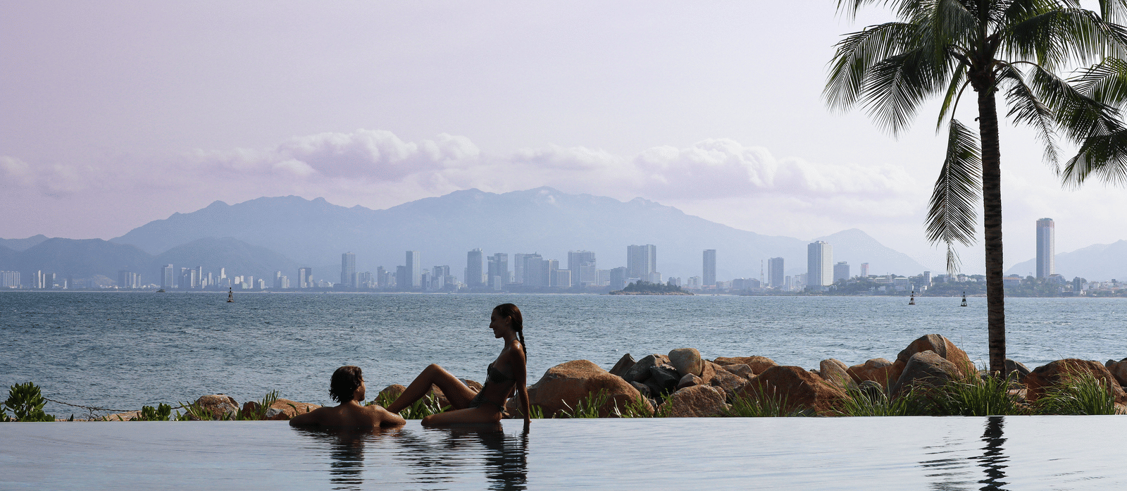 Silhouette of a couple at a swimming pool and beyond it there are some rocks and a palm tree.